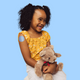 A young girl with curly black hair wearing a yellow floral top and white skirt, holding a brown teddy bear wearing a Thimble Prepare patch against a solid light blue background.